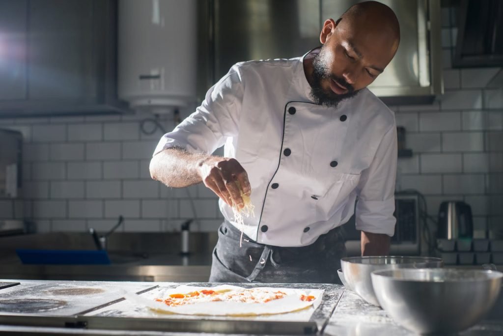 cook-in-kitchen-8629129 A professional chef adds cheese to a pizza in a modern kitchen setting.